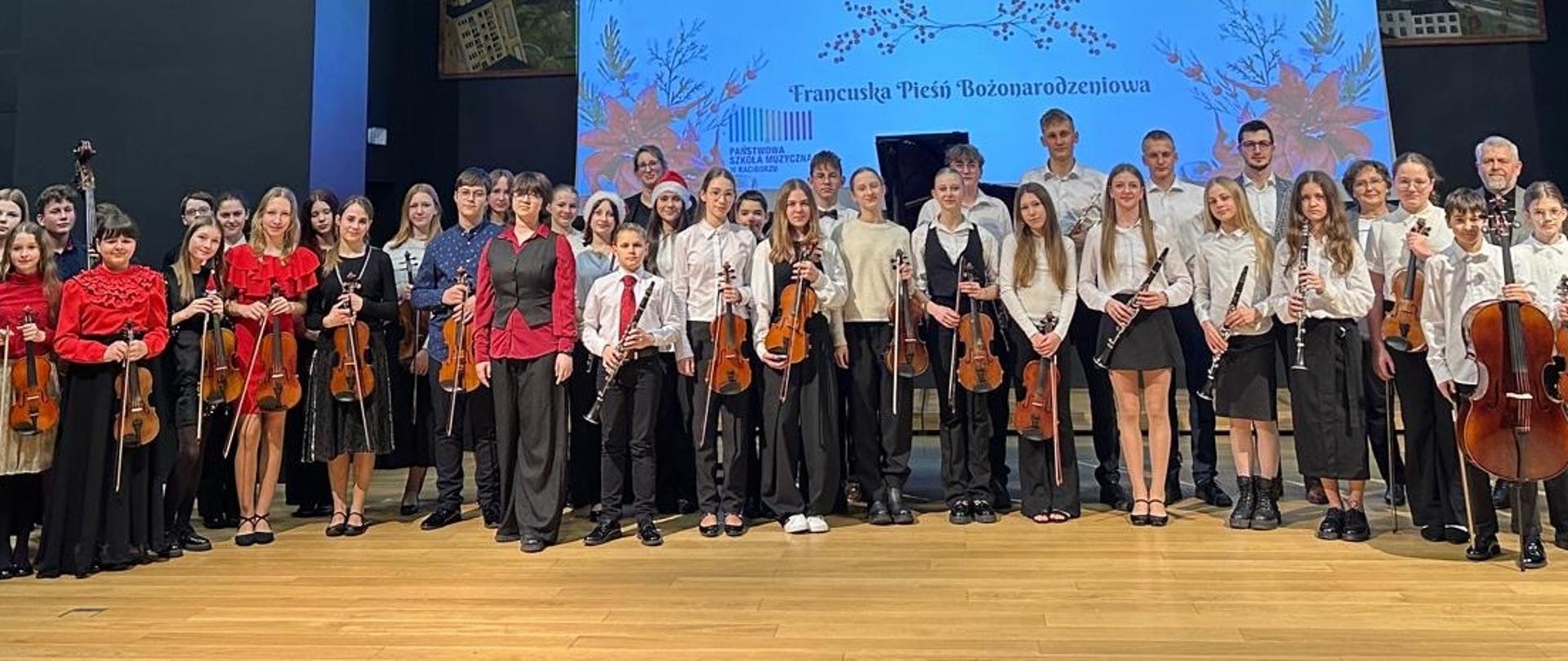 Na zdjęciu widać dużą grupę młodych muzyków stojących na scenie w sali koncertowej. Trzymają instrumenty smyczkowe, takie jak skrzypce, altówki, wiolonczele i kontrabas. Ubrani są elegancko – część osób ma białe koszule i czarne spodnie lub spódnice, inni czerwone bluzki lub kamizelki. W tle znajduje się ekran z prezentacją w świątecznym, bożonarodzeniowym klimacie. Cała grupa pozuje do wspólnego zdjęcia po występie.