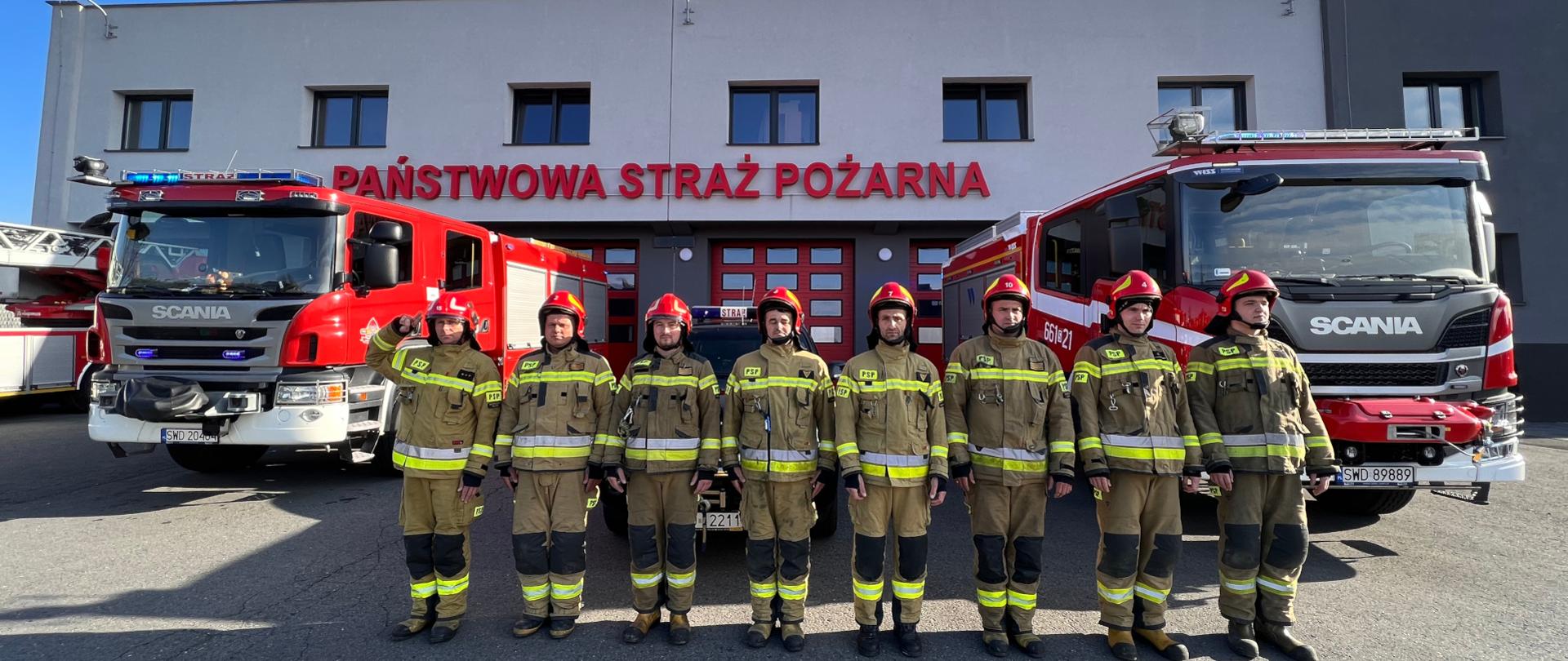Przed garażem Jednostki Ratowniczo-Gaśniczej strażacy ubrani w ubrania specjalne stojąc w szeregu oddają hołd, w tle dwa samochody ratowniczo - gaśnicze
