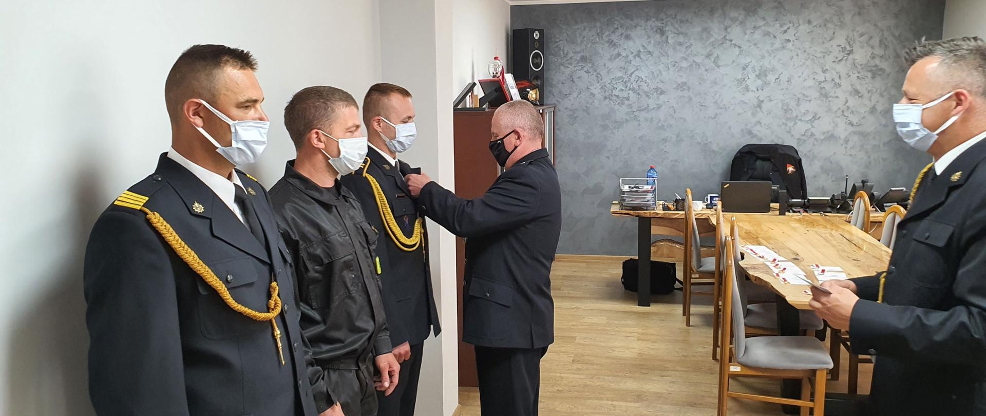 Zdjęcie przedstawia pięciu strażaków w mundurach zebranych w pomieszczeniu. Trzech z nich stoi w rzędzie. Jednemu z nich przypinany jest medal do klapy marynarki. Wszyscy mają założone maseczki ochronne. W tle wyposażenie biurowe gabinetu.