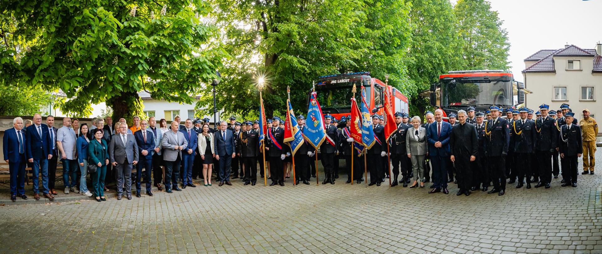 Zdjęcie przedstawia uczestników mszy świętej po jej zakończeniu pozujących do pamiątkowego zdjęcia na tle samochodów ratowniczo-gaśniczych.