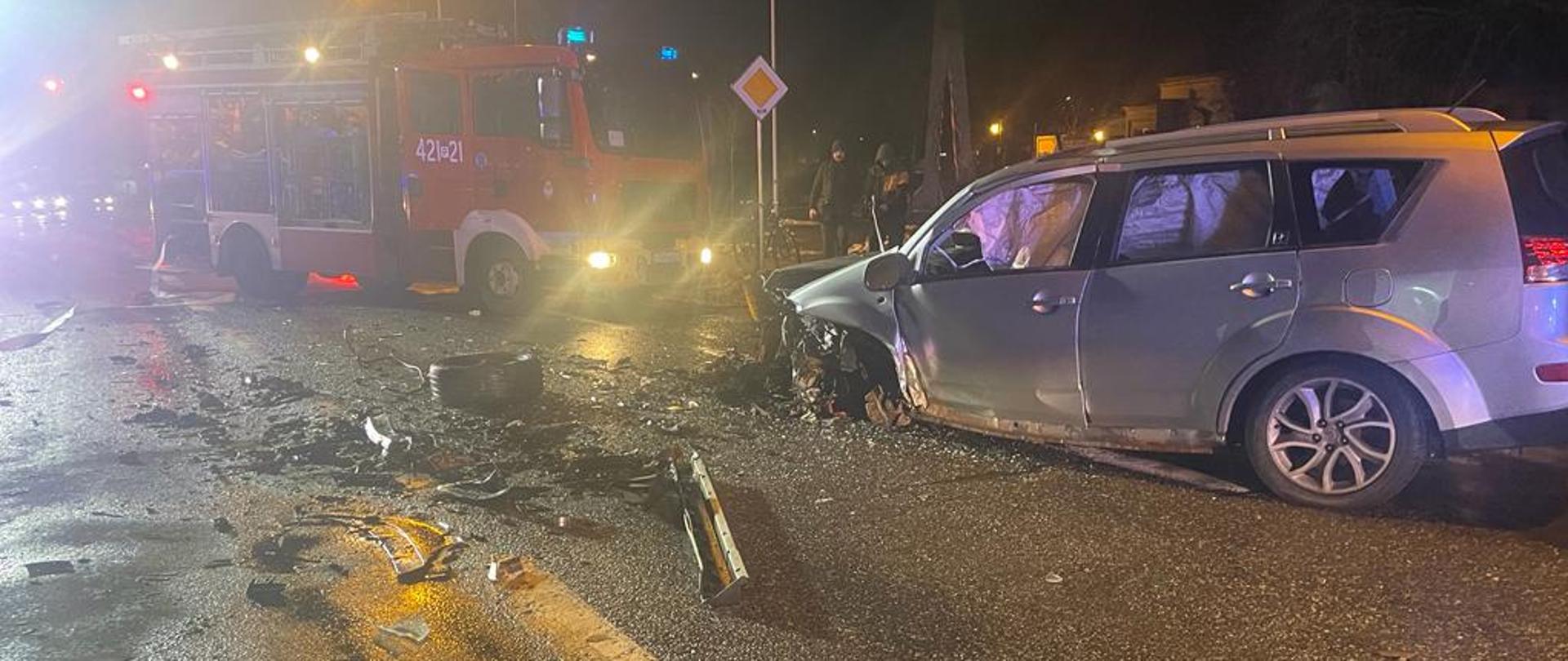 Na zdjęciu widać uszkodzony pojazd znajdujący się na drodze. W tle widać samochód strażacki. Jest pora nocna