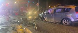 Na zdjęciu widać uszkodzony pojazd znajdujący się na drodze. W tle widać samochód strażacki. Jest pora nocna