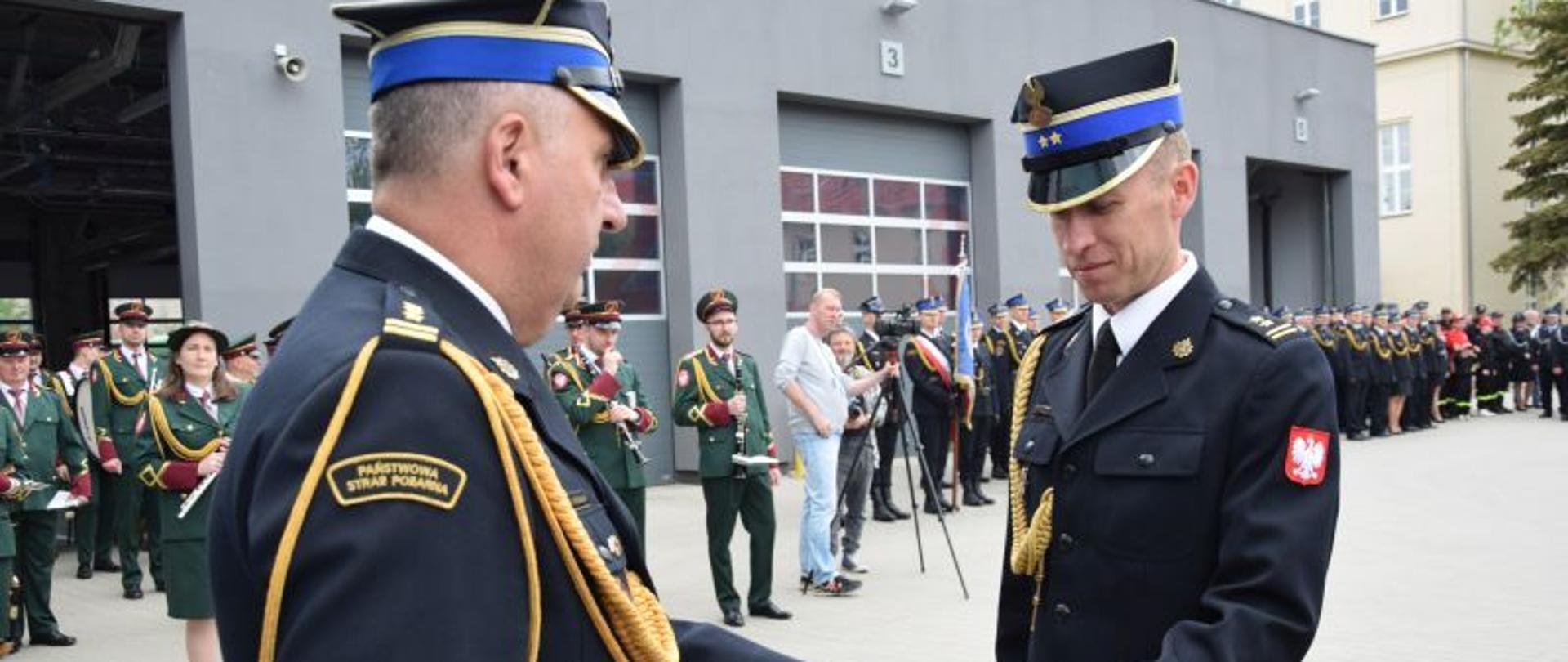 strażak wręcza drugiemu strażakowi statuetkę, w t;e grupa ludzi i szary budynek