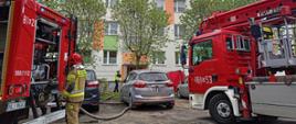 Zdjęcie przedstawia akcję gaśniczą. Strażak obsługujący autopompę na tle budynku wielorodzinnego, w którym wybuchł pożar 