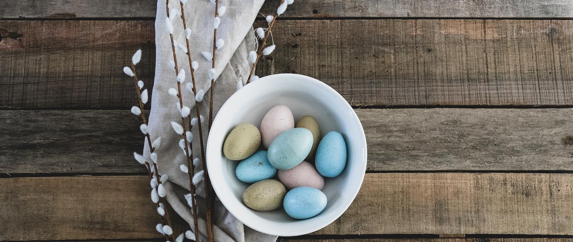 W misce są pisanki. Obok miski leżą gałązki z baziami oraz ściereczka płócienna.