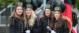 Grupa młodych dziewczyn w galowych górniczych czarnych strojach i czarnych czapkach z pióropuszami stoi na dworze przed schodami, w tle zielone drzewa.