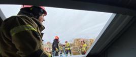 Strażak w hełmie specjalistycznym sprzęcie stoi w trzymając linę zabezpieczając strażaków ćwiczących na barierkach ubranych w specjalistyczny sprzęt wysokościowy oraz hełmy.