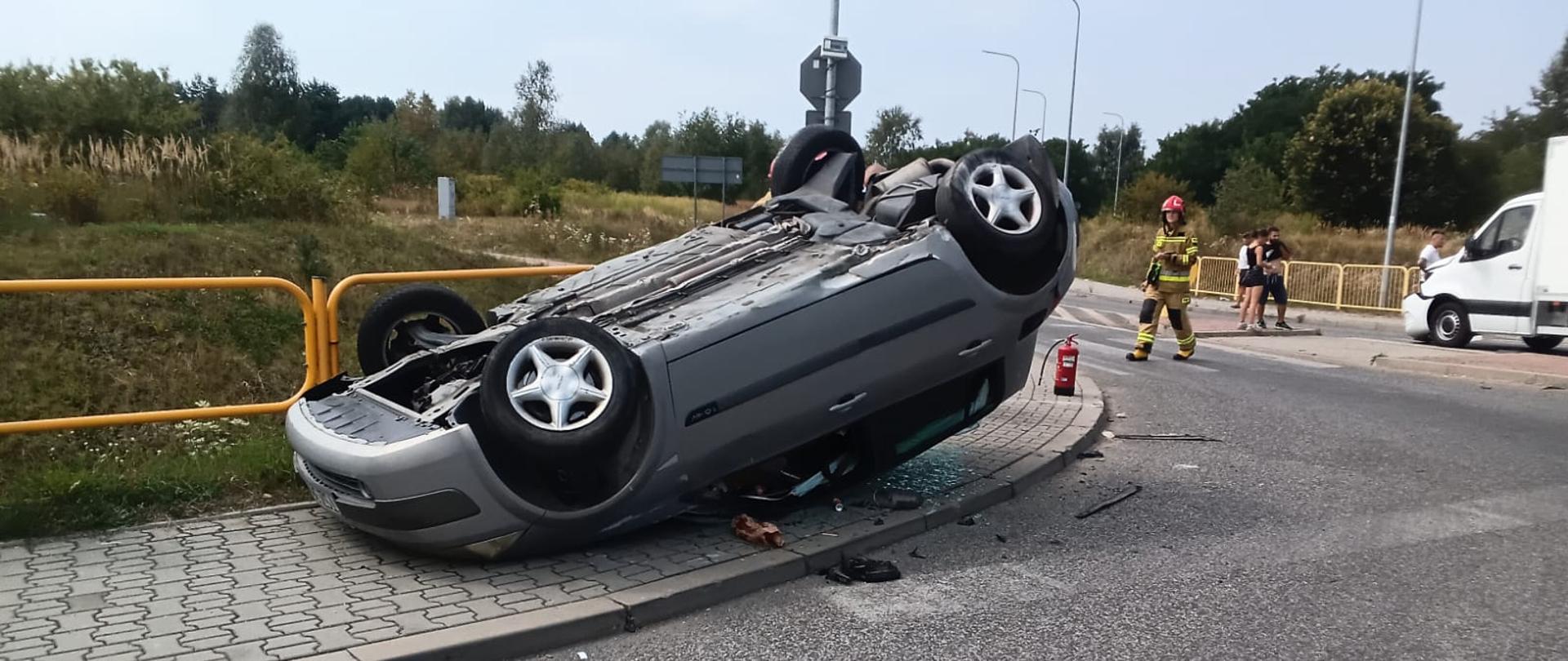Zdjęcie przedstawia miejsce wypadku: w centralnym punkcie na dachu leży samochód osobowy w tle przechodzi ratownik a po prawej stronie widzimy drugi z biorących udział w zdarzeniu pojazdów.