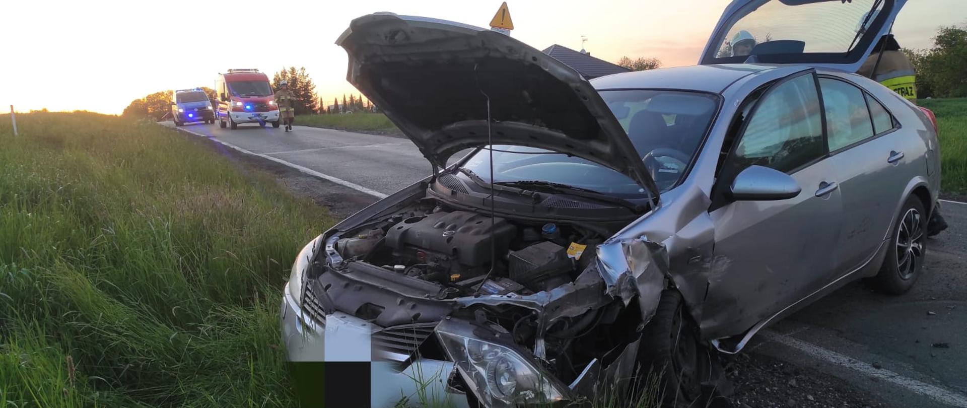 Widać uszkodzony przód samochodu, pojazd jest w rowie i na drodze, w oddali samochód straży pożarnej