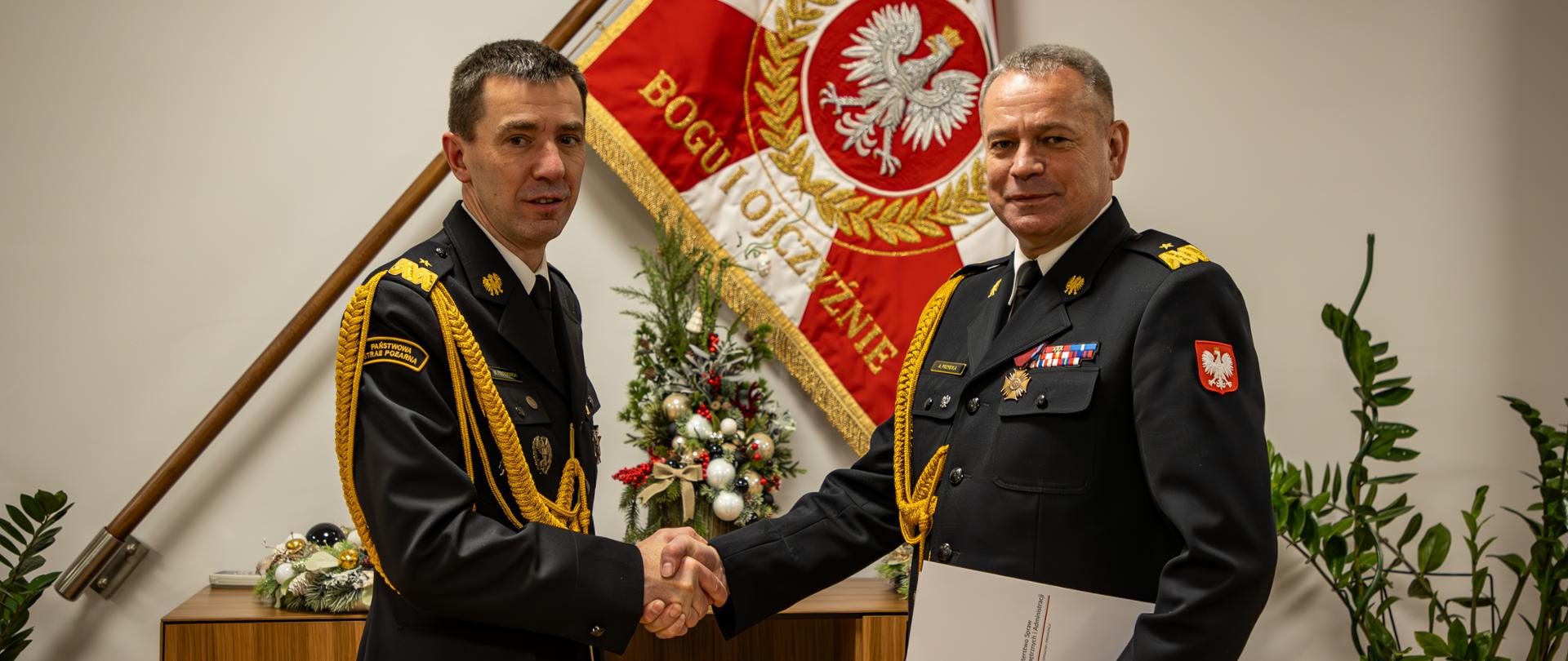 Ubrani w mundury galowe nadbrygadier Mariusz Feltynowski oraz nadbrygadier Arkadiusz Przybyła ściskają dłonie. W tle sztandar PSP oraz dekoracje. 