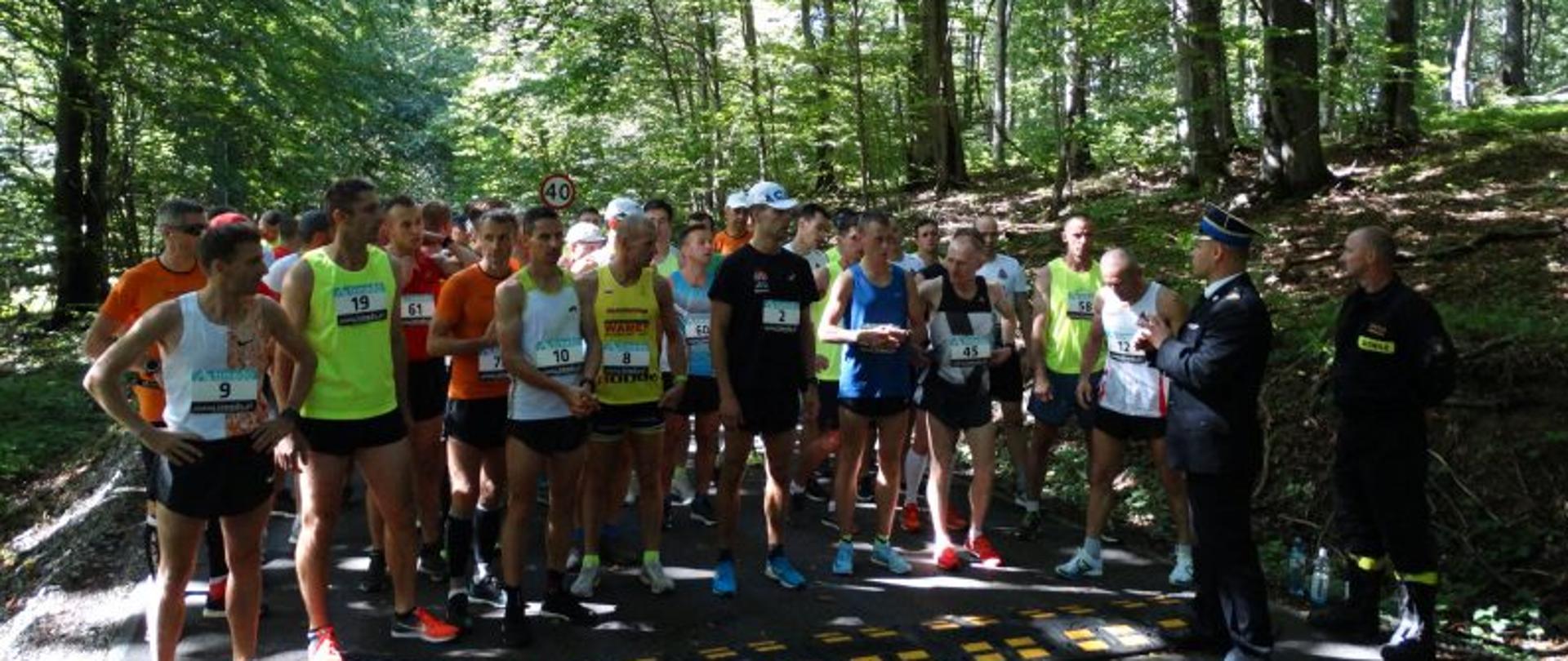 Na zdjęciu las,grupa biegaczy stoi na starcie, obok młodszy brygadier Piotr Śmietana