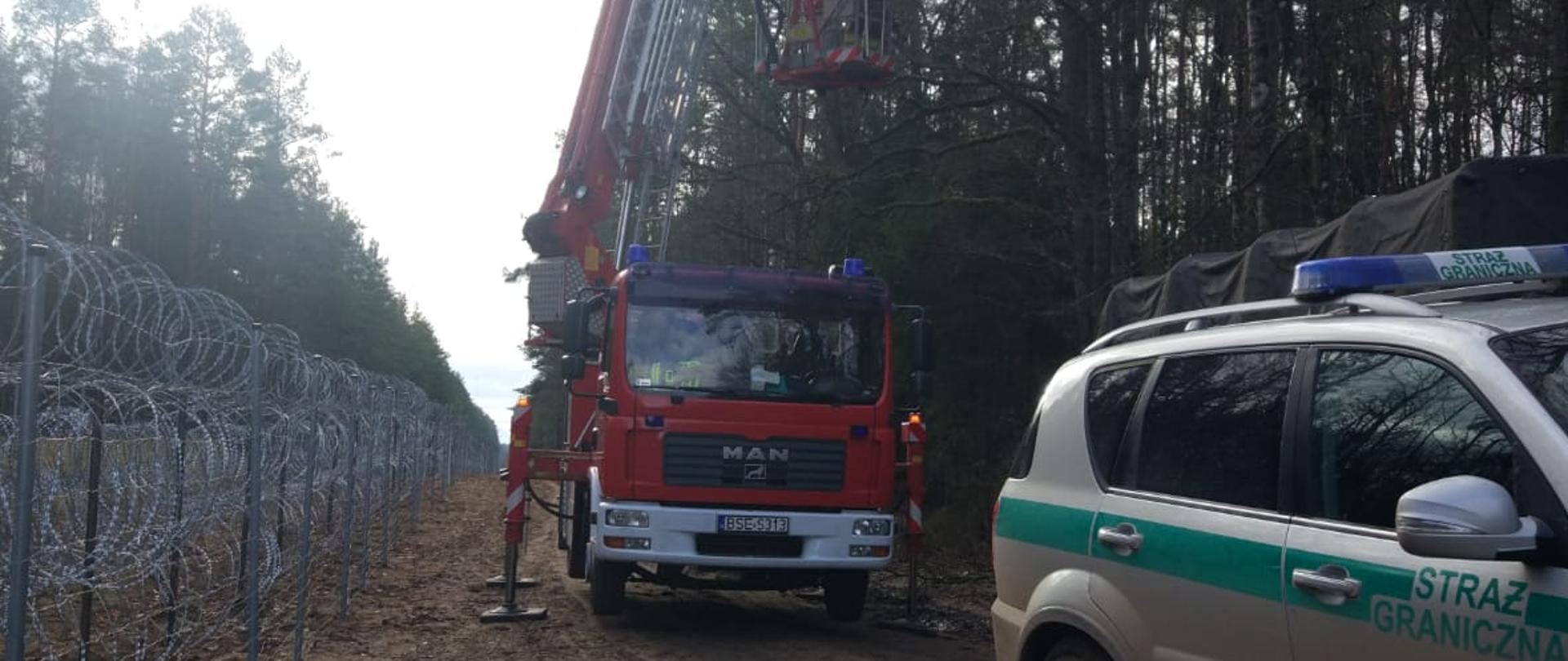 Na granicy przy płocie z kolczastego drutu ustawiony samochód straży granicznej, za nim samochód straży pożarnej - w koszu podnośnika strażak który przycina drzewa połamane silnym wiatrem