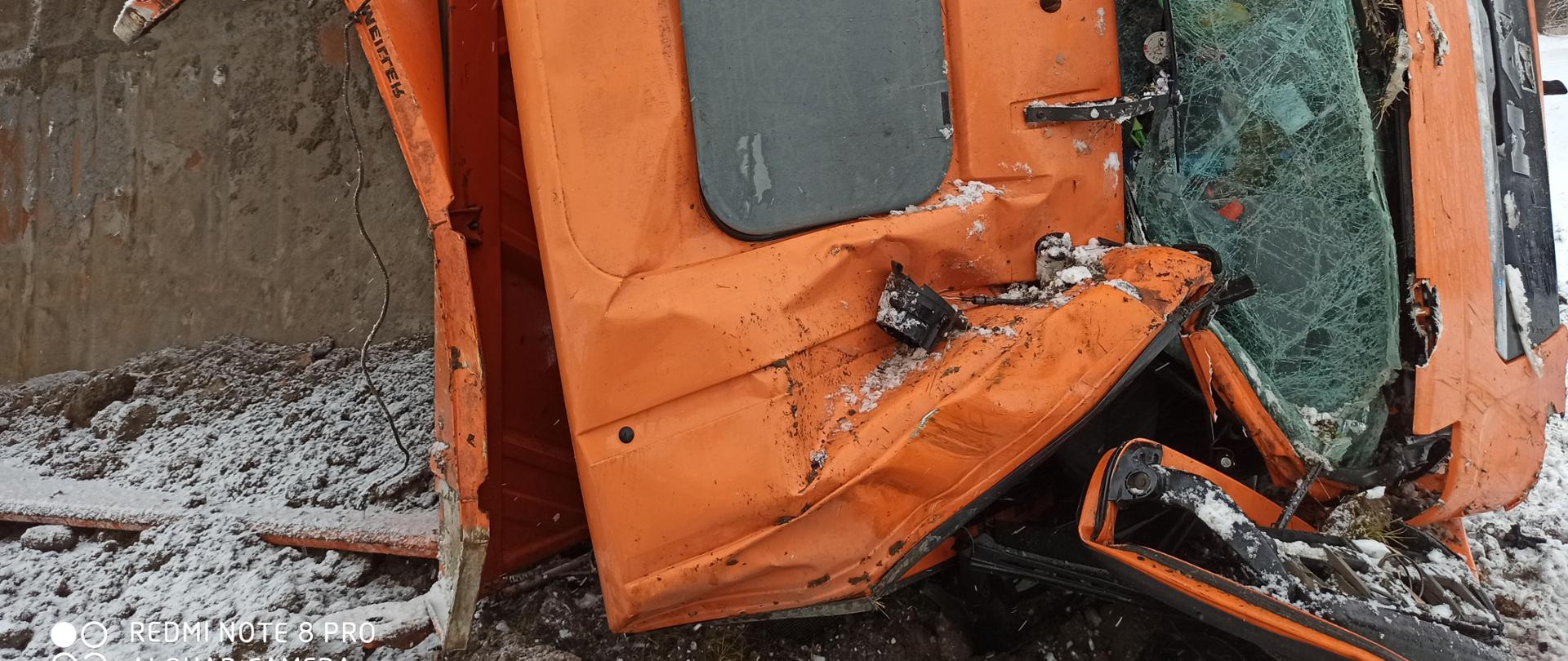 Widok ciężarówki po wypadku. Samochód ciężarowy ze skrzynią ładunkową leży na prawym boku. Kabina zgnieciona w wielu miejscach. Przednia szyba rozbita. Prawe drzwi wyłamane.