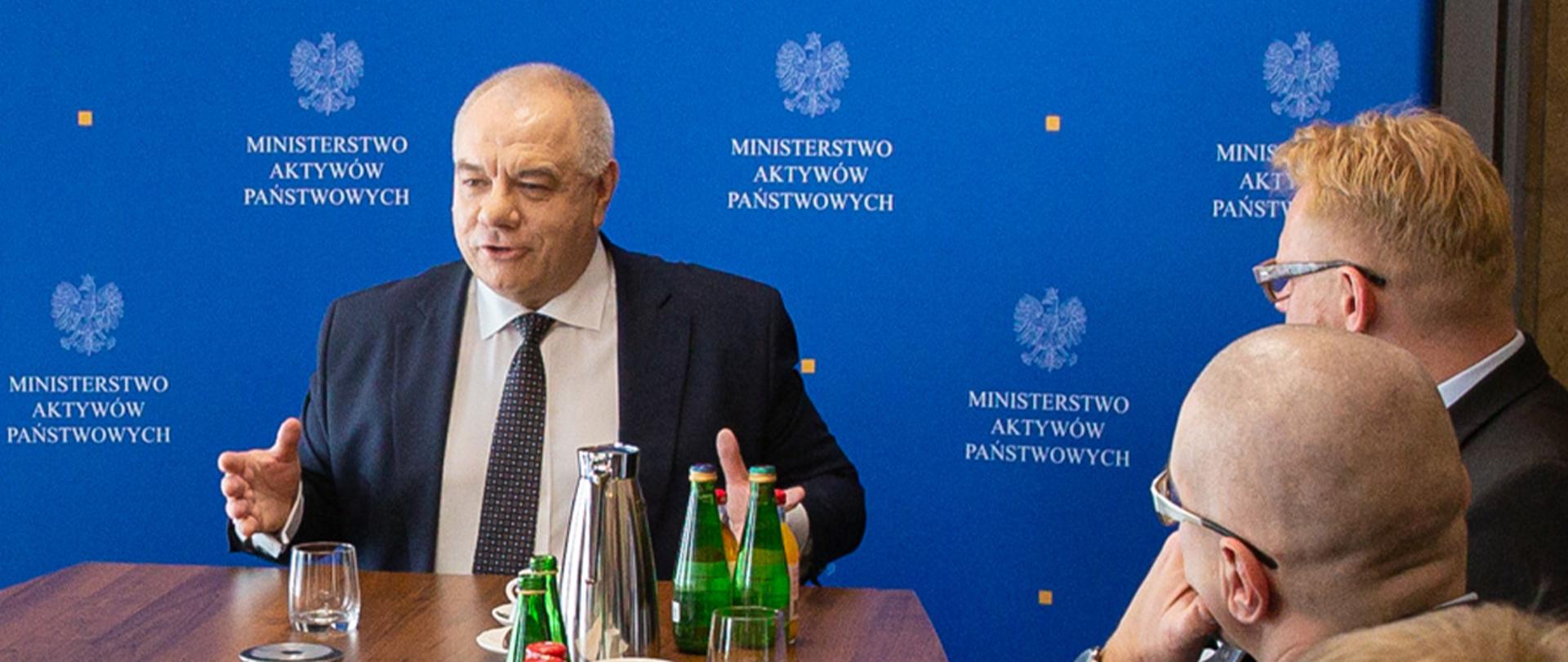 Wicepremier Jacek Sasin i dwóch innych mężczyzn siedzą za stołem. W tle ścianka z logotypami MAP. 