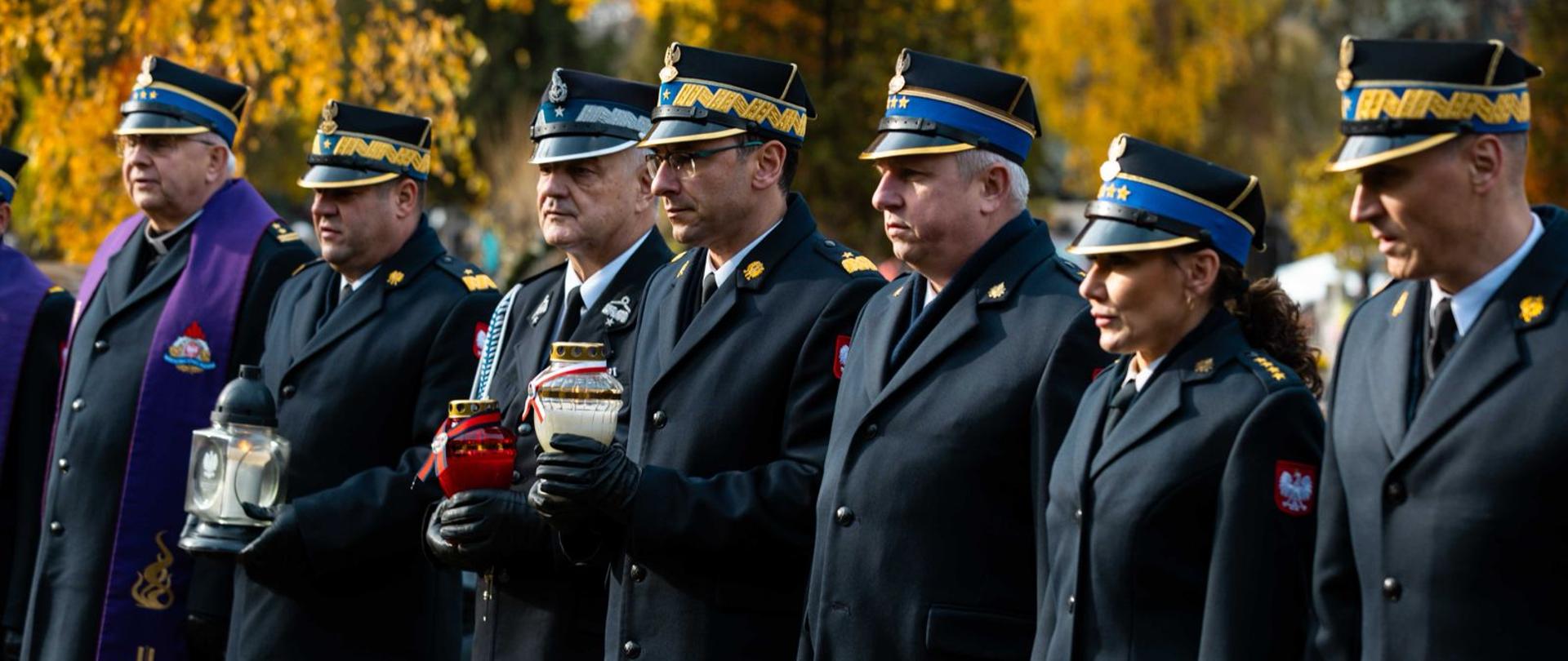 Sześciu mężczyzn i jedna kobieta, wszyscy w mundurach galowych z czapkami na głowach, stoją w rzędzie na zewnątrz. Ubrani są w ciemne, dwurzędowe płaszcze lub kurtki i czapki ze złotymi emblematami i otokami. Większość ma na ramionach złote gwiazdki oznaczające stopień, a niektórzy mają też medale na piersi. Trzy osoby trzymają w rękach znicze w pojemnikach (jeden z fioletową, a dwa z czerwoną oprawą). Mężczyzna najbardziej po lewej ma na sobie fioletową stułę, co sugeruje, że jest duchownym.
Tłem jest park lub cmentarz z drzewami o jesiennych, złoto-brązowych liściach i rozmyty blask jesiennego słońca.
