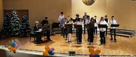 Zdjęcie przedstawia uczniów i nauczycieli szkoły grających na instrumentach na scenie na sali koncertowej. Z przodu stoją cztery uczennice grające na fletach. Za nimi stoją uczniowie grający na saksofonach i klarnecie. Za nimi stoi nauczyciel grający na saksofonie oraz nauczyciel grający na trąbce. Po lewej stronie przy pianinie siedzi uczeń, który akompaniuje zespołowi oraz uczeń grający na perkusji. Po lewej stronie sceny stoją trzy udekorowane choinki. Przód sceny ozdobiony jest światełkami i balonami w kolorach: pomarańczowy, fioletowy i niebieski. 