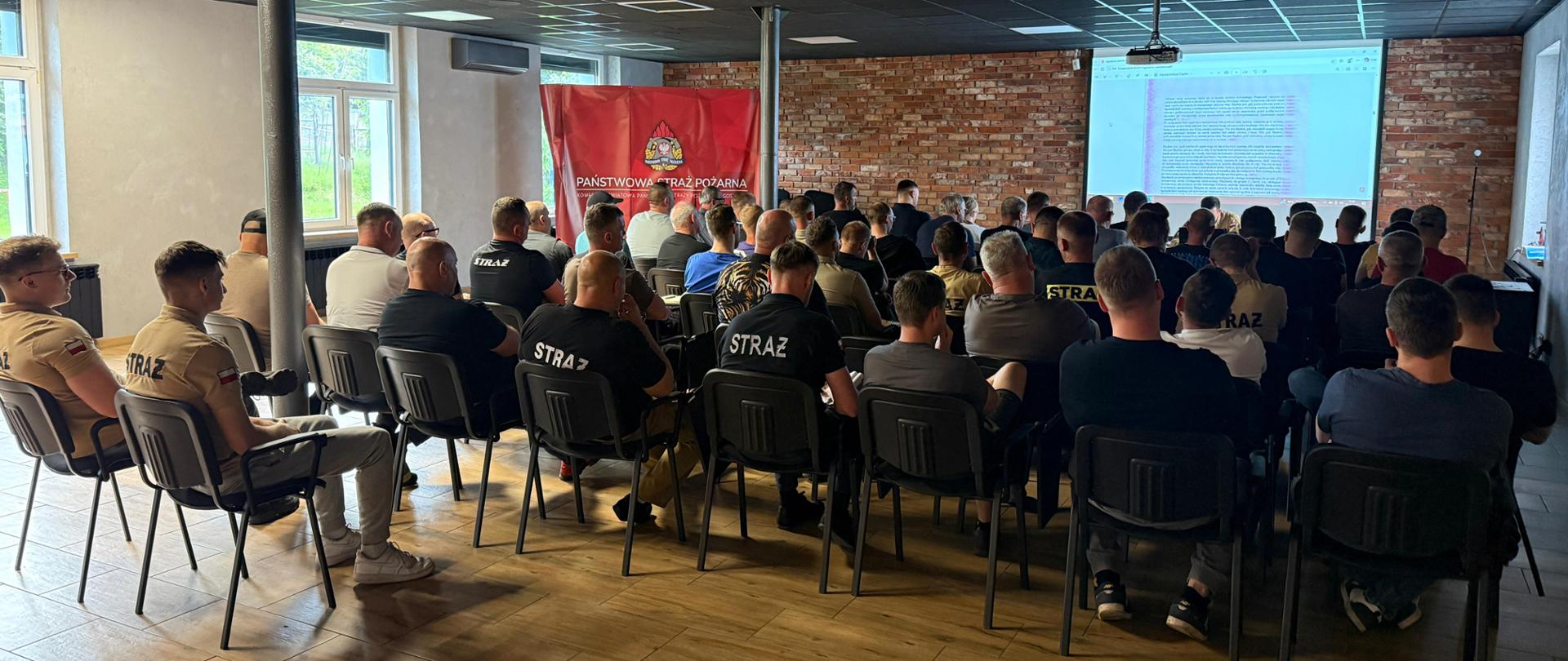 Druhów, którzy siedzą w sali na szkoleniu. Patrzą oni na wyświetlaną na rzutniku prezentacje. Szkolenie prowadzi strażak.