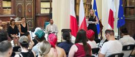 Pani Ambasador Małgorzata Kopeć przemawiająca podczas Narodowego Czytania w Bibliotece Narodowej Malty. W tle flagi. Na zdjęciu widać również część osób czytających oraz widownię.