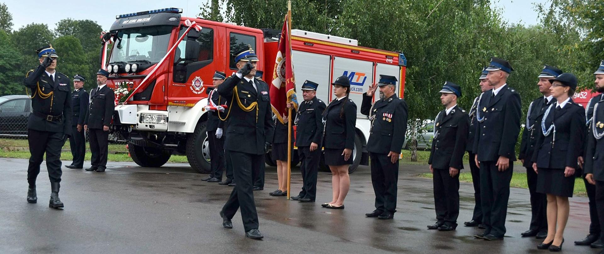 Komendant wojewódzki PSP, na prawo stoją ustawieni w rzędzie ubrani w mundury. W tle samochód pożarniczy i drzewa