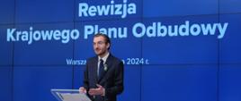 Wiceminister funduszy i polityki regionalnej Jan Szyszko, minister stoi przy pulpicie z mikrofonem, za jej plecami grafika z niebieskim tłem i informacją o rewizji KPO 