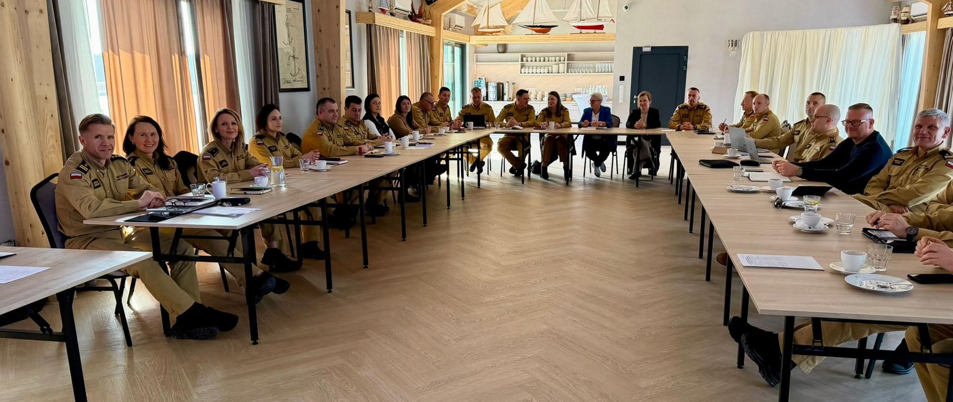 Szerokie ujęcie wnętrza przestronnej sali konferencyjnej o wystroju marynistycznym, ze stromym sufitem z widocznymi drewnianymi belkami. Dwa długie stoły są ustawione w kształcie litery V, a przy nich siedzą uczestnicy spotkania. Widać strażaków w piaskowych mundurach służbowych oraz osoby cywilne. W tle, na półkach, ustawione są liczne modele żaglowców. Uczestnicy są zwróceni w kierunku centralnego punktu spotkania.