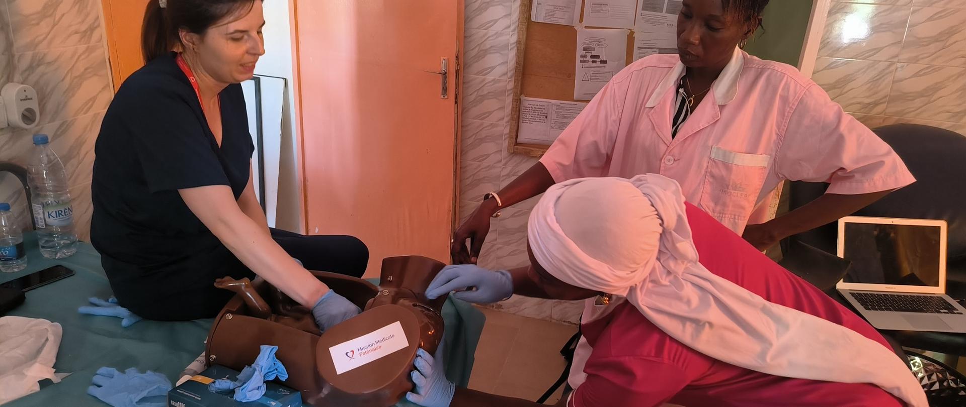 Trzy kobiety w strojach pracowników medycznych. Dwie wykonują ćwiczenia na fantomie, trzecia się przygląda