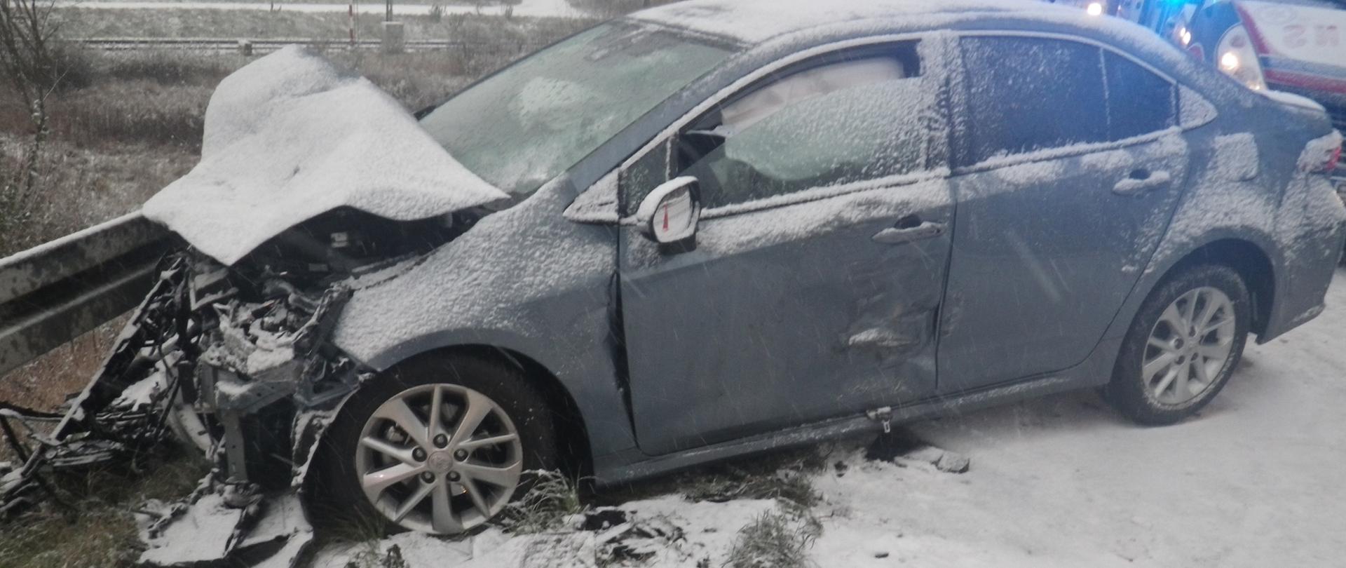 Na środku zdjęcia widoczny jest samochód osobowy Toyota Corolla koloru niebieskiego, który w wyniku zdarzenia zatrzymał się przodem na barierze energochłonnej. Cała przednia część pojazdu jest zniszczona zaś maska w wyniku zdarzenia została pogięta i wypiętrzyła się do góry. Pojazd ma także wgniecione i porysowane przednie drzwi od strony kierowcy. W pojeździe w drzwiach kierowcy jest lekko uchylona szyba, za którą widoczne są wystrzelone kurtyny boczne. Tył pojazdu nie uległ zniszczeniu i stoi na drodze asfaltowej pokrytej warstwą śniegu. Przed pojazdem zarówno na drodze jak i na porośniętym trawą poboczu widoczne są odłamane elementy uszkodzonego pojazdu. Za samochodem w lewym górnym rogu zdjęcia widoczne są tory oraz znajdujące się w oddali zabudowania. W prawym górnym rogu zdjęcia widoczne są stojące za uszkodzonym samochodem pojazdy służb ratowniczych. Pierwszym z pojazdów jest samochód Zespołu Ratownictwa Medycznego koloru białego z czerwonymi i niebieskimi pasami, za którym znajdują się dwa samochody ratowniczo-gaśnicze koloru czerwonego należące do Straży. W oddali za pojazdami służb widoczne są przejeżdżające samochody osobowe. 