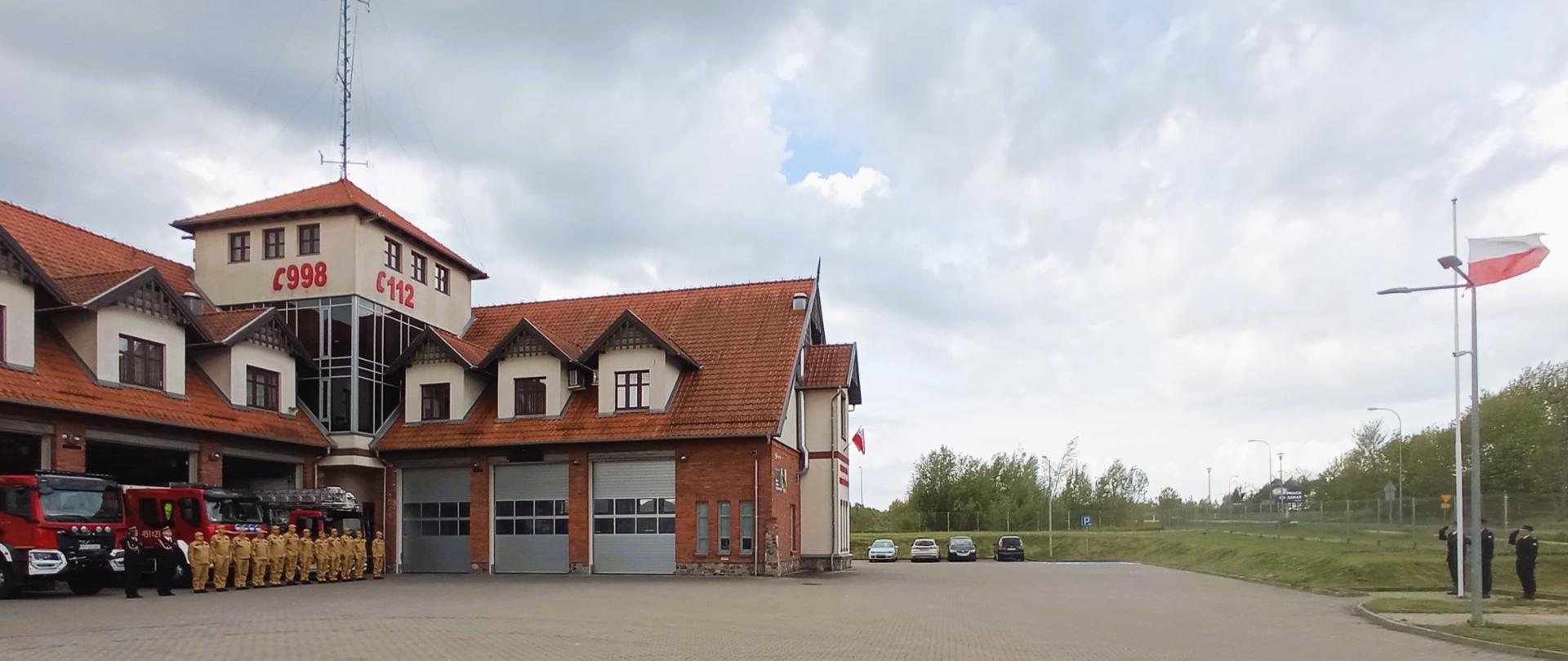 Zdjęcie przedstawia strażaków Komendy Powiatowej Państwowej Straży Pożarnej w Gołdapi podczas podnoszenia flagi Rzeczypospolitej Polskiej przy akompaniamencie hymnu państwowego z okazji Dnia Flagi Państwowej Rzeczypospolitej Polskiej. Pierwszy z lewej w pododdziale st. bryg. Wojciech Szczepanik Komendant Powiatowy Państwowej Straży Pożarnej w Gołdapi oddaje honor przez salutowanie. Funkcjonariusze usytuowani są na tle pojazdów pożarniczych częściowo zlokalizowanych w obiekcie strażnicy. Z prawej strony widać strażaków wchodzących w skład pocztu flagowego, podczas podnoszenia flagi Rzeczypospolitej Polskiej