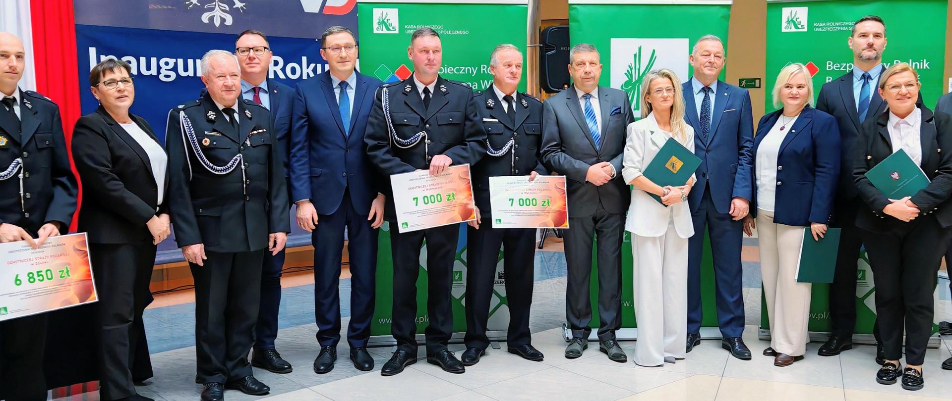 Kilkanaście osób stojących w szeregu podczas oficjalnego wydarzenia. W centrum znajdują się mężczyźni w mundurach OSP trzymają w dłoniach duże czeki i dokumenty. Po bokach stoją osoby w eleganckich strojach – kobiety i mężczyźni. W tle widać banery kolorowe banery KRUS