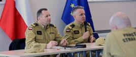 Na zdjęciu widać od lewej zastępcę komendanta głównego PSP nadbryg. Adama Koniecznego oraz komendanta głównego PSP nadbryg. Andrzeja Bartkowiaka, którzy siedzą przy stoliku przemawiając do zebranej kadry. w tle flagi Polski i Unii Europejskiej oraz prezentacja.