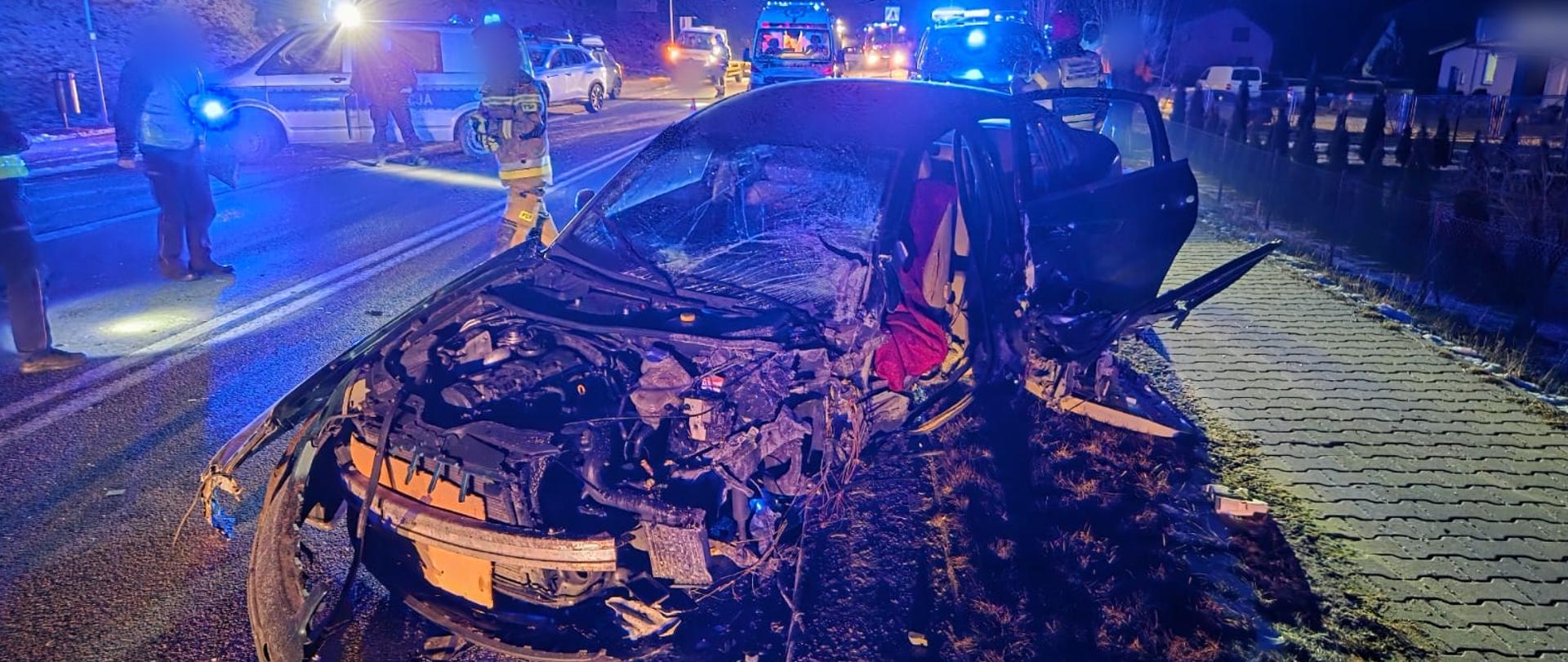 Samochód zniszczony blokuje drogę, w tle strażak