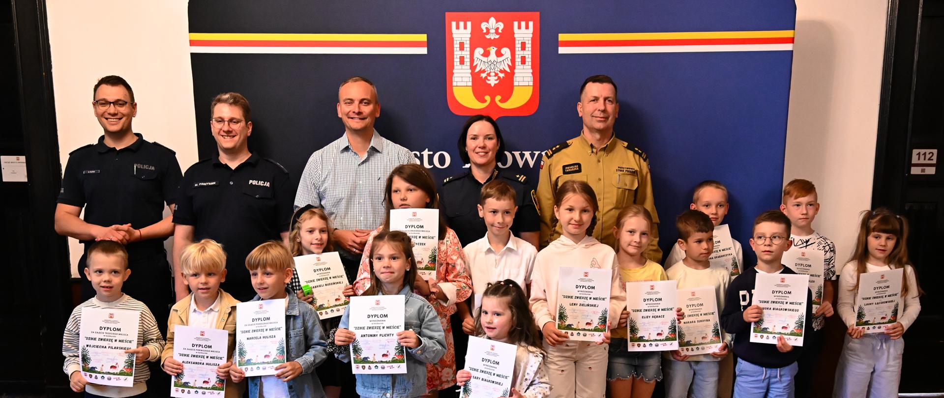 Prezydent Inowrocławia Arkadiusz Fajok wraz z funkcjonariuszami Inowrocławskiej Policji, Straży Miejskiej, Zastępcą Komendanta Powiatowego Państwowej Straży Pożarnej bryg. Ireneuszem Taraszką i Laureatami konkursu „Dzikie zwierzę w mieście” tuż po wręczeniu dyplomów uczestnikom konkursu