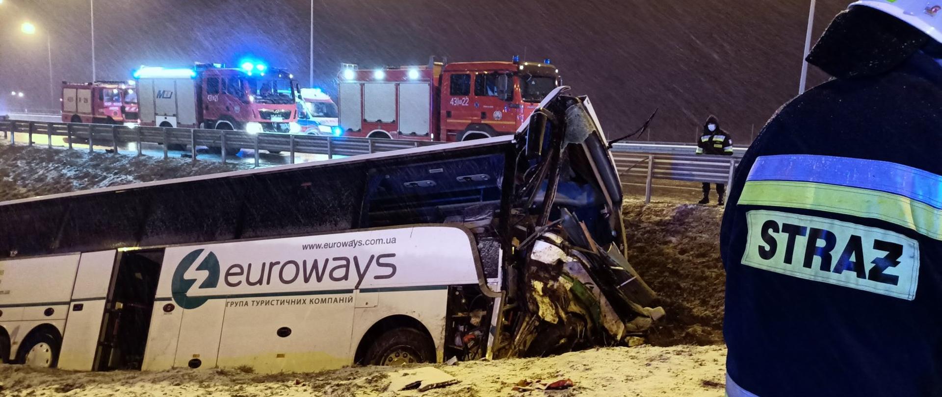 Zdjęcie zrobione w nocy. Na pierwszym planie po lewej stronie autokar z rozbitym przodem. Pojazd jest poza jezdną. Po prawej stronie widać strażaka w ubraniu specjalnym z napisem STRAŻ na plecach. W tle widać trzy samochody pożarnicze.