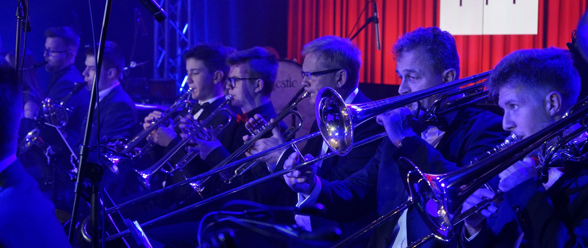 zdjęcie zawiera sekcję instrumentów dętych blaszanych i drewnianych podczas wykonywania koncertu. Od lewej dwie osoby grające na saksofonach, trzy na trąbkach, dwie na puzonach w niebieskiej poświacie światła scenicznego. W tle widoczne elementy sceny oraz telebim z czerwoną scenografią.