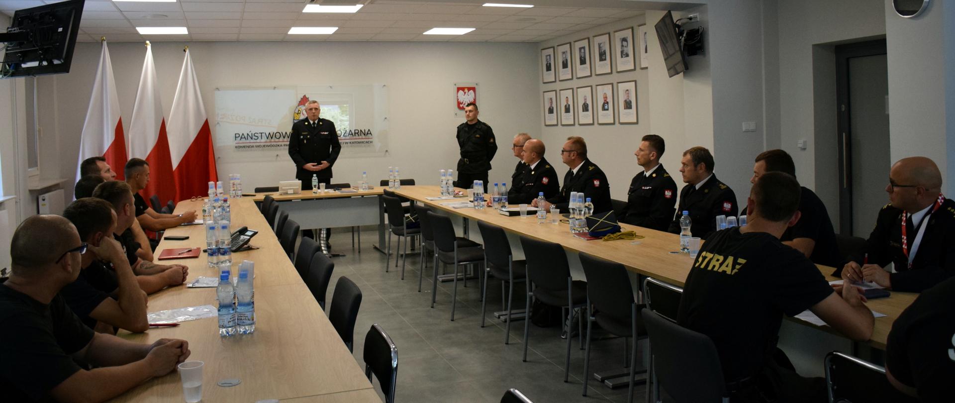 Na zdjęciu widoczna jest sala sztabowa w Komendzie Wojewódzkiej PSP w Katowicach. Po obydwu stronach za stołami siedzą w mundurach strażacy ratownicy OSP i komendanci powiatowi i miejscy PSP. Za stołem stoi zastępca śląskiego komendanta wojewódzkiego PSP st. bryg. Rafał Świerczek , który przemawia.