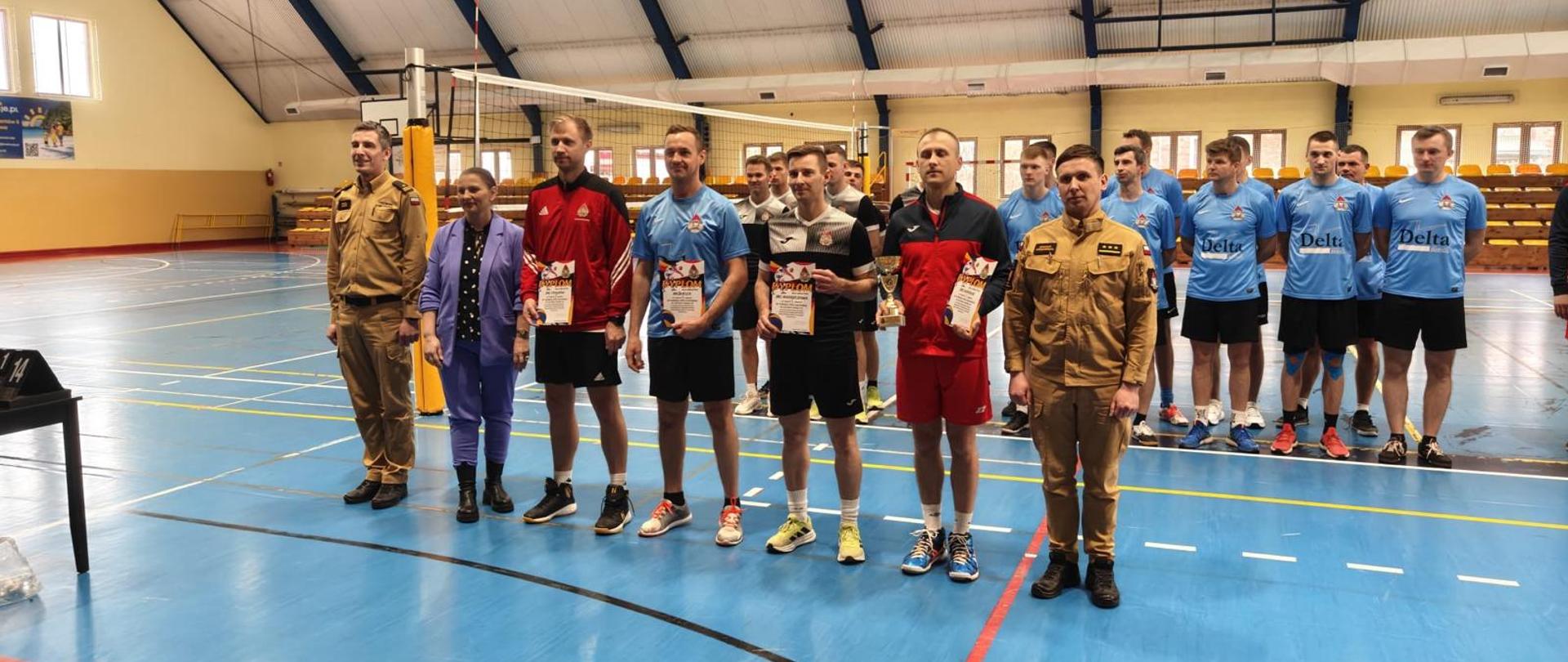 Sala w obiekcie w którym przeprowadzono rozgrywki w piłkę siatkową. W rzędzie stoją kapitanowie drużyn biorących udział w zmaganiach, wraz z zastępcą Komendanta Powiatowego Państwowej Straży Pożarnej we Włoszczowie oraz dowódcą Jednostki Ratowniczo-Gaśniczej we Włoszczowie. Zawodnicy w rękach trzymają dyplomy oraz puchar. W tle pozostali zawodnicy.