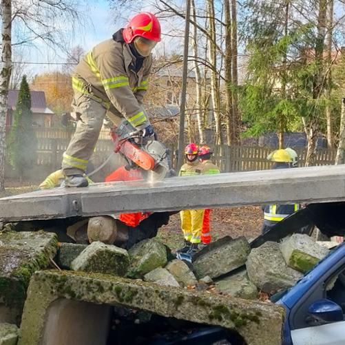 Strażak przecina piłą mechaniczną betonowy kloc, z tyłu widać innych strażaków oraz domy w tle
