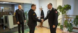 Opolski Komendant Wojewódzki PSP st. bryg. Krzysztof Kędryk w obecności Zastępcy Opolskiego Komendanta Wojewódzkiego PSP st. bryg. Dariusza Gieronia wręcza powierzenie obowiązków naczelnika wydziału planowania operacyjnego KW PSP w Opolu st. kpt. Romanowi Wyszce. Strażacy w mundurach wyjściowych. W tle widoczny sekretariat KW PSP Opole.
