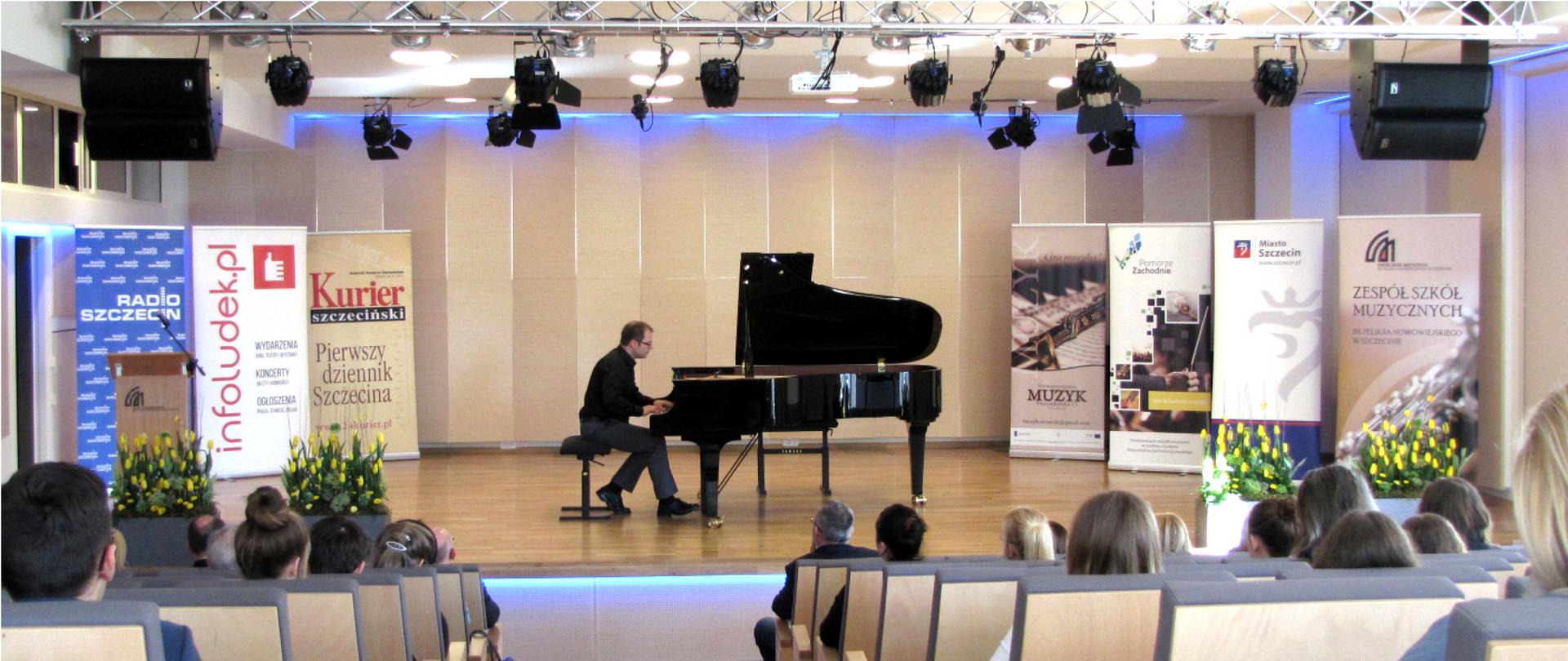 Grafika przedstawia osobę grająca na fortepianie w Sali Koncertowej Szkoły