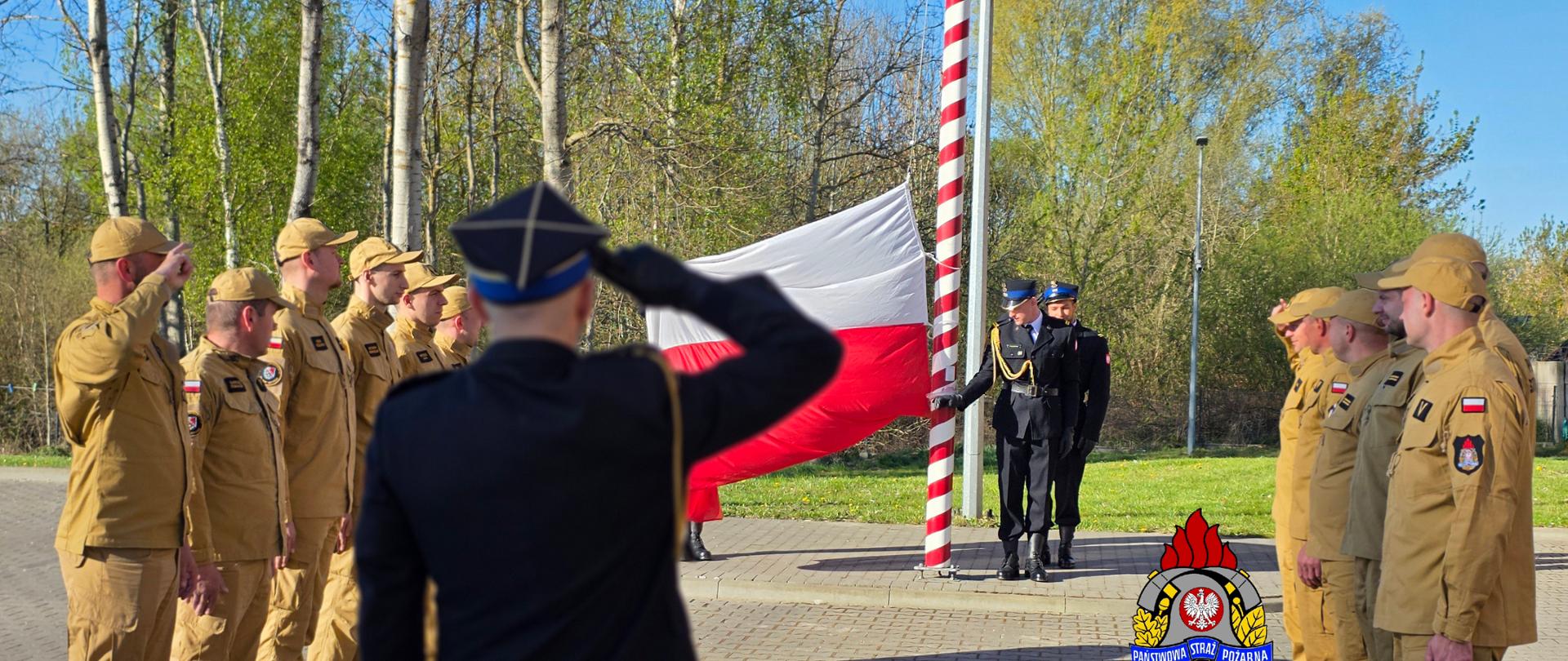Uroczystość podniesienia flagi: strażacy w galowych, czarnych mundurach stoją przy czerwono-białym maszcie i rozwijają biało-czerwoną flagę. Po obu stronach ustawieni są strażacy w beżowych mundurach, stojący w szyku i oddający salut. Na pierwszym planie, tyłem do kamery, widoczna jest osoba w czarnym mundurze salutująca. W tle znajdują się drzewa i trawnik, a na dole widnieje emblemat Państwowej Straży Pożarnej z napisem „Gryfice”.
