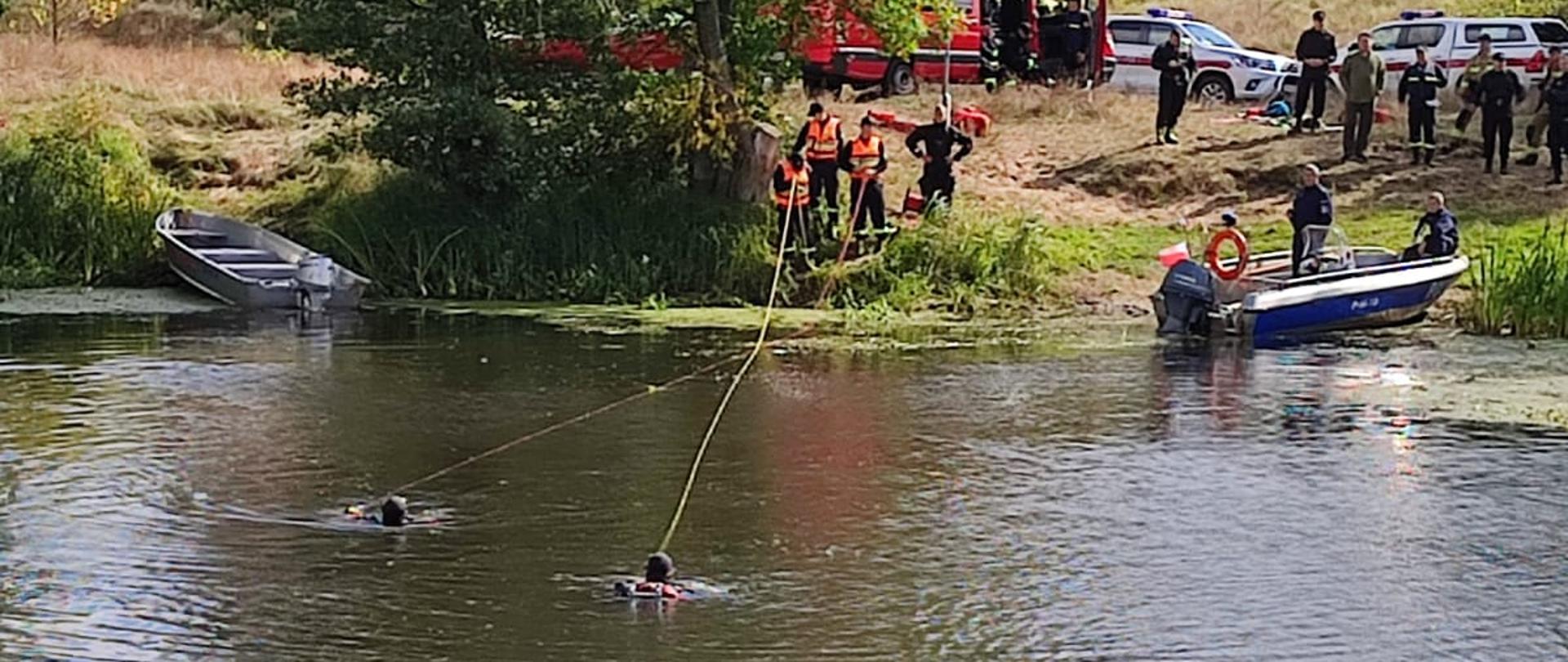 Płetwonurkowie pływają w rzece. Z brzegu zabezpieczają ich linkami strażacy. Dookoła pojazdy straży pożarnej oraz strażacy.