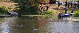 Płetwonurkowie pływają w rzece. Z brzegu zabezpieczają ich linkami strażacy. Dookoła pojazdy straży pożarnej oraz strażacy.