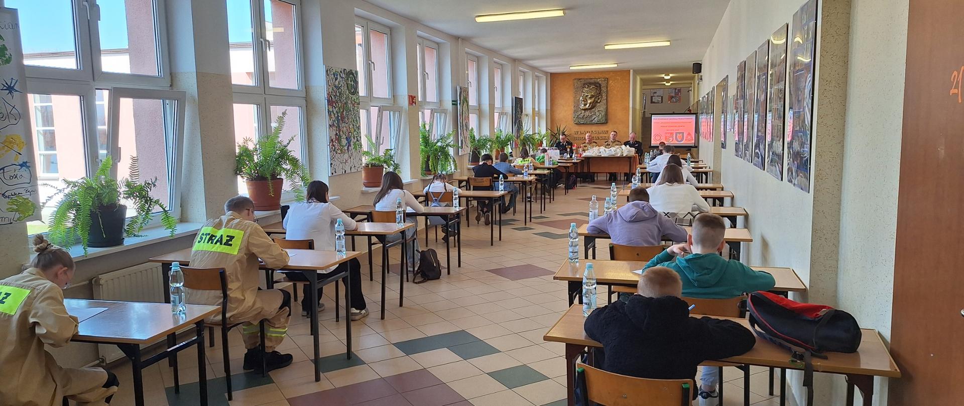 Turniej Wiedzy Pożarniczej. Dzieci siedzą w ławkach i piszą testy. Komisja siedzi przy biurku. 