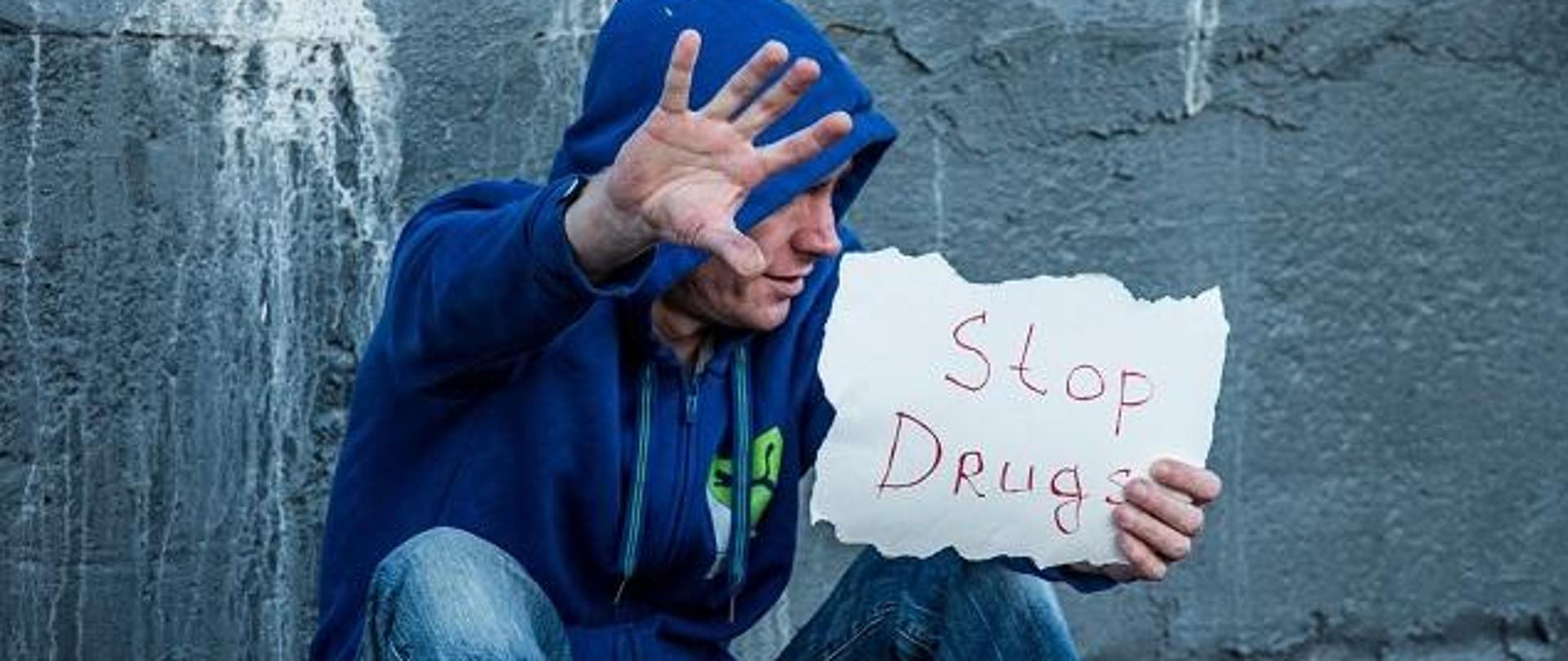 na zdjęciu widać mężczyznę siedzącego na ziemi, w bluzie sportowej, z kapturem na głowie, trzymającego w ręce kartkę z napisem STOP DRUGS