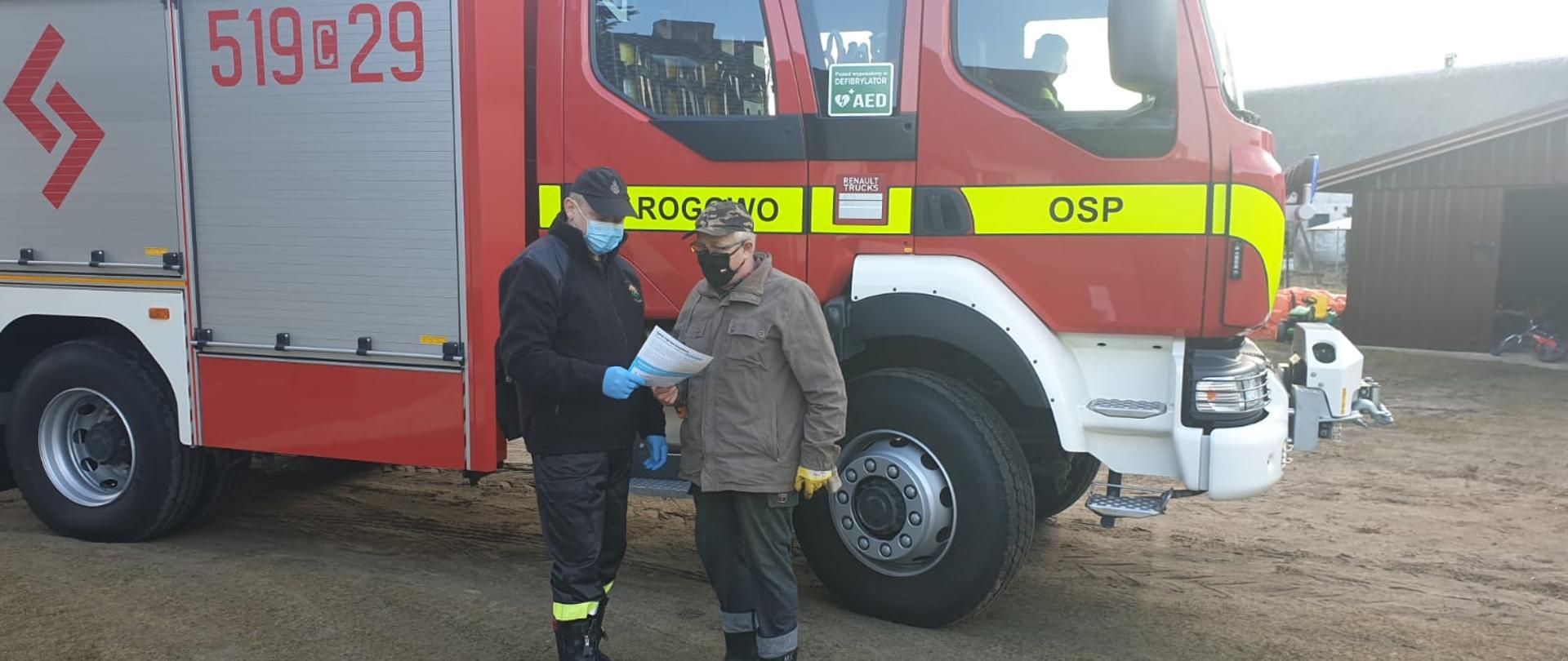 Zdjęcie przedstawia strażaka OSP, który wręcza mężczyźnie ulotkę dotyczącą szczepień przeciwko COVID-19. Na tle mężczyzn znajduje się samochód pożarniczy. Jest to prawa strona pojazdu. Na kabinie pojazdu znajduje się napis Rogowo OSP oraz numery operacyjne 519c29. Za kierownicą w pojeździe widać strażaka kierowcę. W oddali zdjęcia widać budynek gospodarczy. 