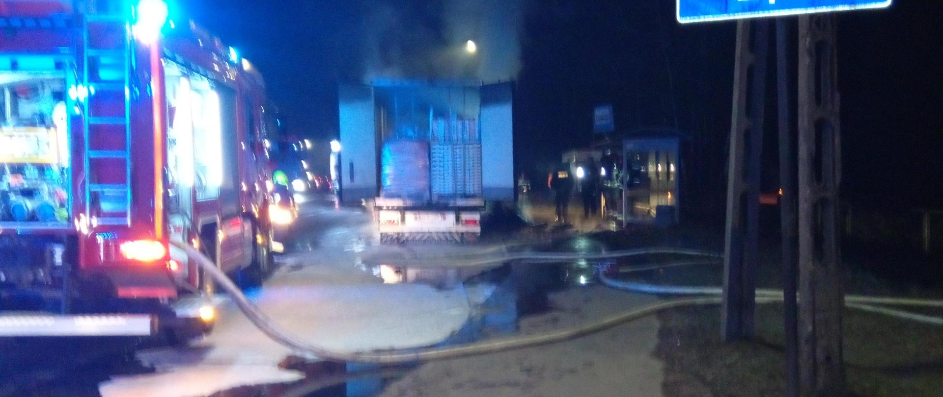 Zdjęcie przedstawia drogę krajową gdzie doszło do pożaru. W centralnym punkcie stoi naczepa z warzywami, po lewej stronie samochód gaśniczy a z prawej zatoczka autobusowa.