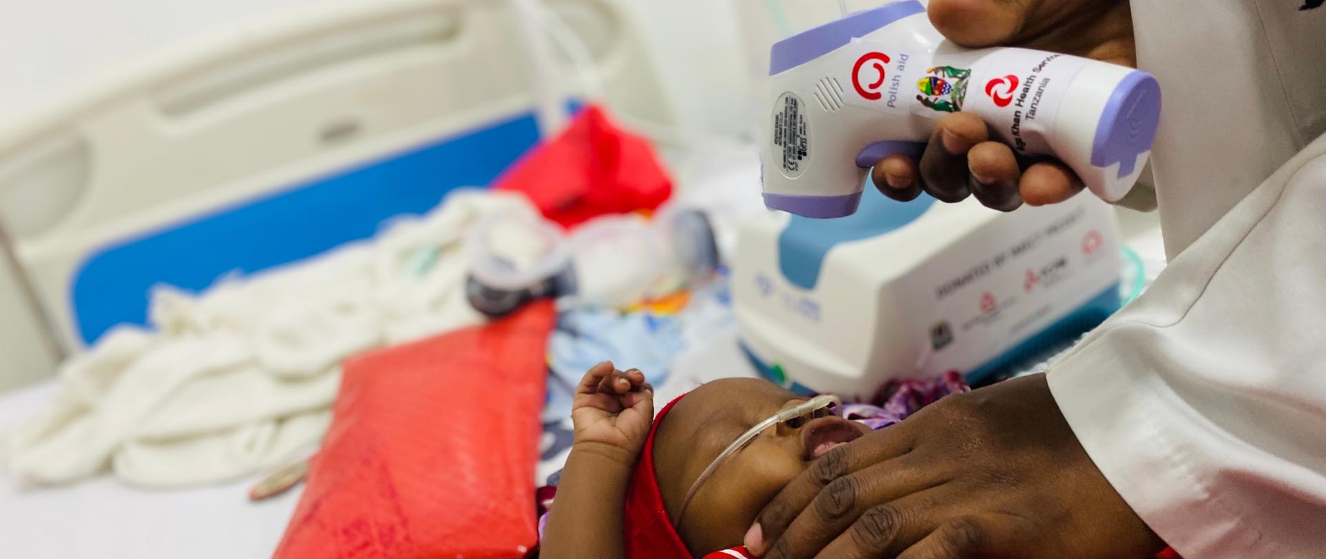 Pracownik medyczny mierzy leżącemu w łóżeczku niemowlakowi temperaturę za pomocą termometru bezkontaktowego