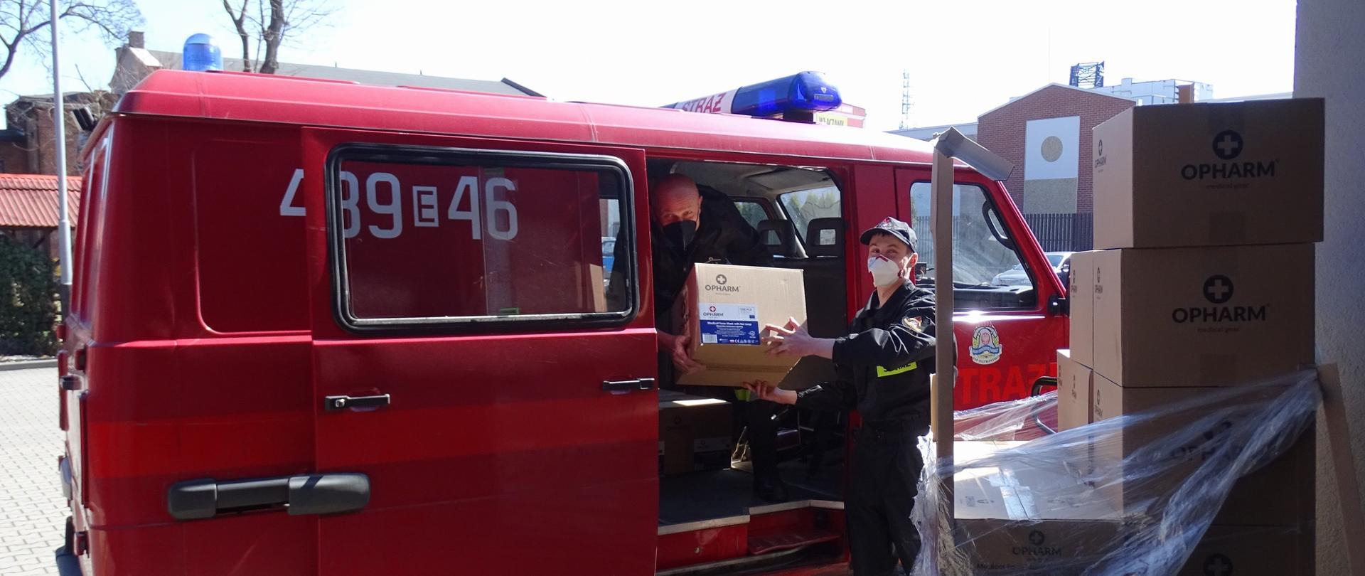 Na zdjęciu strażacy OSP Piątkowisko pakujący środki ochrony indywidualnej do pojazdu ich jednostki. Zdjęcie wykonane na placu wewnętrznym Komendy Powiatowej Państwowej Straży Pożarnej w Pabianicach. Strażacy w umundurowaniu koszarowym. Wszyscy w maseczkach ochronnych na twarzach.