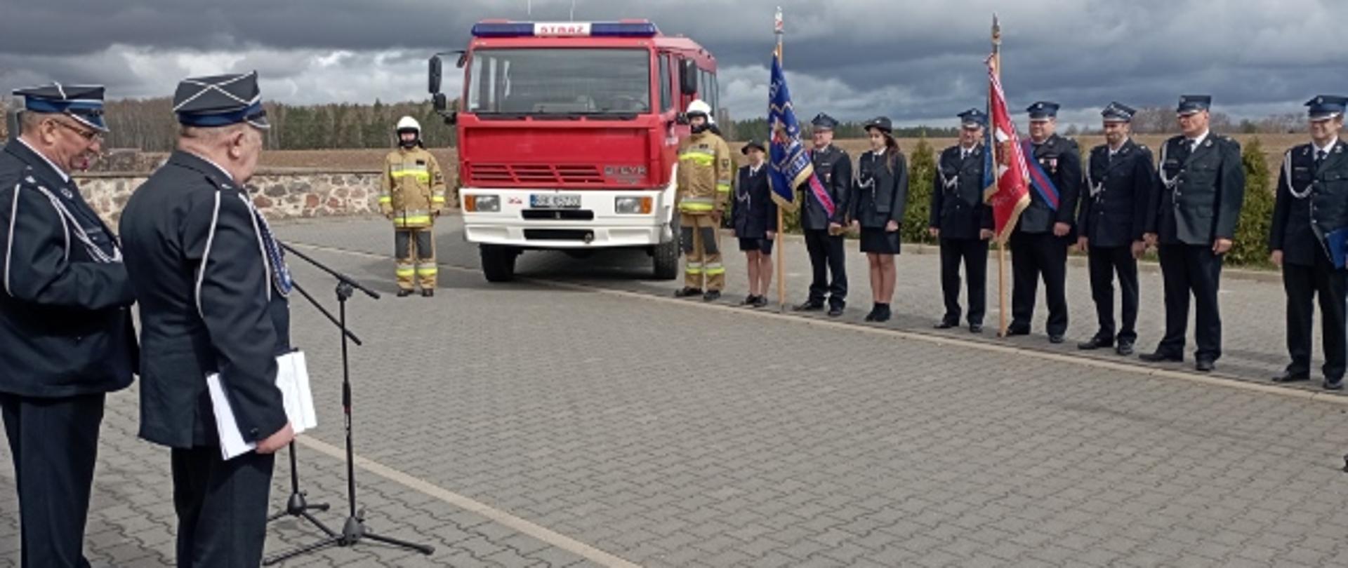 Strażacy i poczty sztandarowe stoją przy nowym wozie 