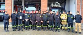 Zdjęcie przedstawia uczestników szkolenia kierowcy-konserwatora sprzętu ratowniczego Ochotniczych Straży Pożarnych, realizowanego w Komedzie Powiatowej Państwowej Straży Pożarnej w Gołdapi, którzy wspólnie z komisją egzaminacyjną, stoją na tle pojazdów pożarniczych zlokalizowanych w garażach strażnicy